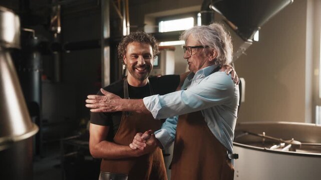 Two men in brown aprons smiling and shaking hands in coffee roasting facility. Standing near industrial roasting machine and celebrating successful discussion or partnership in bright workspace.
