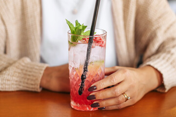 Woman hand holding glass with pink cocktail and ice cubes on wooden table. Fresh drink with mint leaves and fruits served with straw. Refreshing beverage for summer party, cafe or bar lifestyle scene.