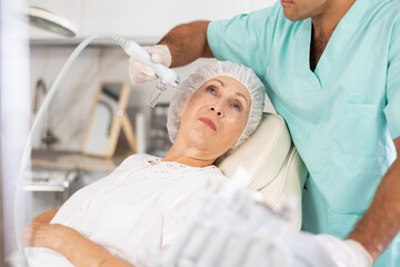 Elderly woman getting facial deep hydration procedure from experienced cosmetologist in aesthetic...