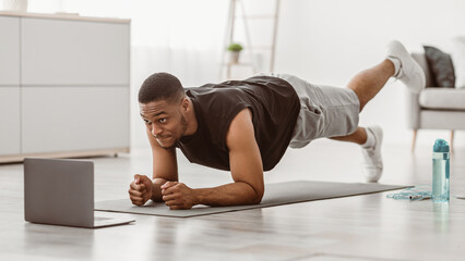 Online Workout. African American Fitness Man Exercising At Laptop Standing In Plank And Doing Leg...