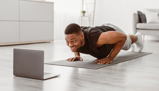 Athletic African American Guy Exercising At Laptop Doing Push-Ups In Living Room At Home. Male Workout Routine And Bodybuilding Concept. Modern Sporty Lifestyle. Selective Focus - Powered by Adobe