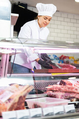 Smiling young female butcher in white uniform and rubber gloves working at counter, slicing fresh...