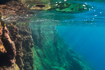 Over-under photograph of a majestic submerged rock wall with sunlight on the surface and a deep...