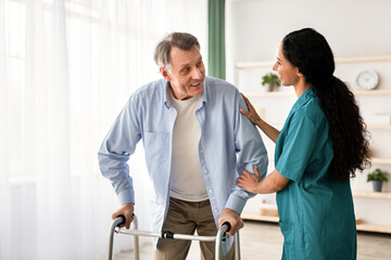 Young nurse helping older man to walk with frame at retirement home. Mature male petient with...