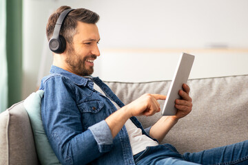 Positive bearded man with headset and digital tablet reclining on couch and smiling, home interior,...