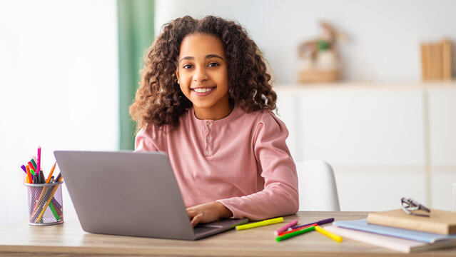 E-Learning. Cheerful african american girl studying on laptop computer learning at home, doing her homework and browsing the internet. Schoolgirl sitting at desk and smiling to camera - Powered by Adobe