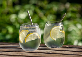 glasses of water with ice and lemon on wooden table in green sunny summer garden