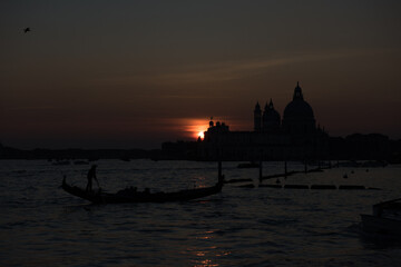 Obraz premium A gondolier navigates a gondola on the water in Venice, Italy, as the sun sets behind the Basilica di Santa Maria della Salute.