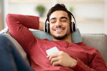 Joyful arab guy listening to music at home, reclining on couch with closed eyes and smiling, using...