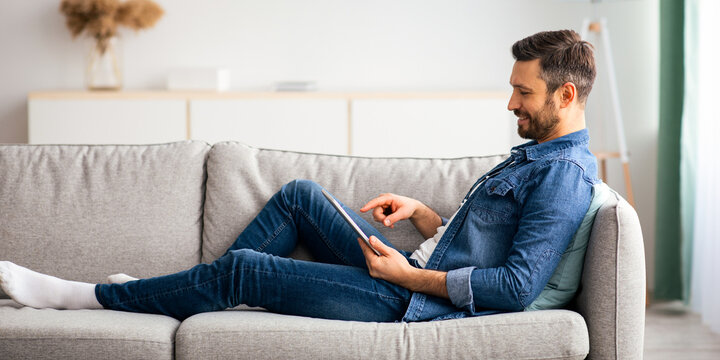Relaxed bearded man using digital tablet while resting at home, side view, copy space. Happy middle-aged man reclining on couch in living room, websurfing or checking social media