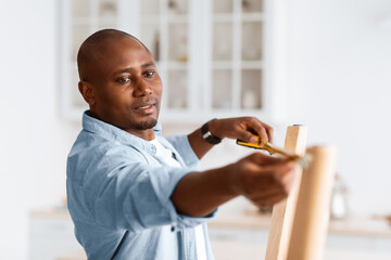 African american handyman working at home, measuring the width between legs of wooden table....