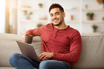 Relaxed arab man sitting on couch at home, using laptop and smiling, drinking coffee and watching...