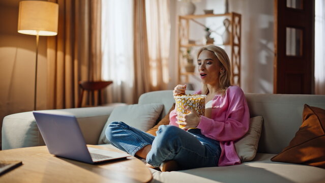 Cheerful blonde streaming series sitcom on laptop snacking chips in comfy house