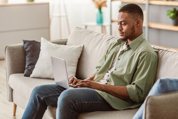 Freelance Career. Serious Black Man Using Laptop Browsing Internet Sitting On Couch At Home. Side...