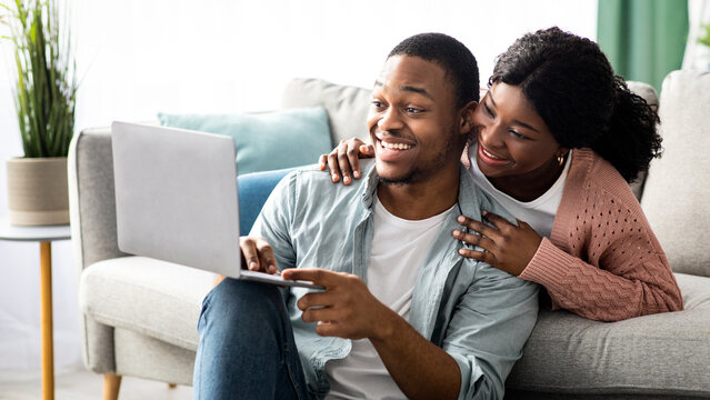 Happy young black couple shopping online at home, using laptop, copy space. Emotional african american guy showing his girlfriend something exciting on Internet, holding notebook - Powered by Adobe