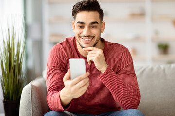 Positive arab guy using brand new mobile phone at home, having online party with friends, using...