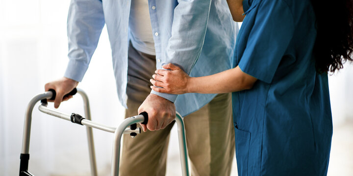 Cropped view of elderly man using walker, young nurse supporting and assisting him at retirement home, closeup. Senior male moving around with walking frame, medical worker supporting him