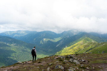 Solo backpacker contemplating expansive mountain panorama with multiple green ridges and forest...