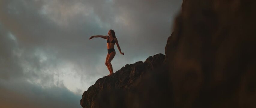 Woman strikes expressive dance pose on cliffside during dramatic dusk