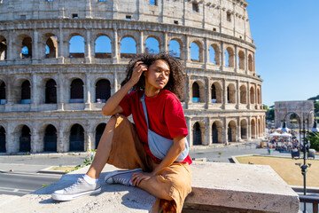 A stylish young woman elegantly sits on a ledge, gracefully posing while showcasing the iconic Colosseum in Rome, framed beautifully against a clear and vibrant blue sky that enhances the scene