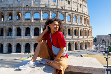 A stylish young woman in a vibrant red shirt and brown pants enjoys a sunny day at Romes Colosseum, blending rich culture with contemporary fashion, enhancing the lively atmosphere
