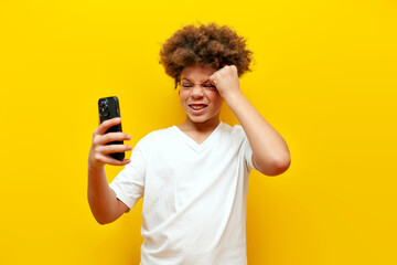 tired unhappy african american boy using smartphone and showing facepalm on yellow isolated...