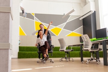 office workers in business suits having fun and riding on chairs with raised hands, african...