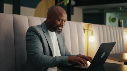 Smiling mature black businessman in blazer using laptop in office lounge - Powered by Adobe