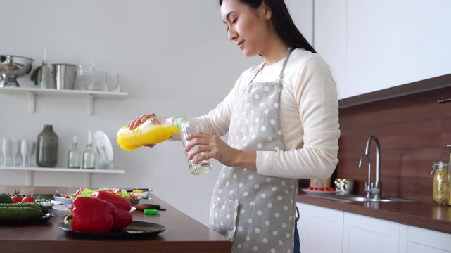 young asian brunette woman in apron pouring orange juice from bottle into glass, korean girl holding and drinking fruity yellow drink in kitchen at home