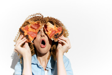 Young woman with curly hair playfully holding pizza slices up to her face, expressing surprise and...