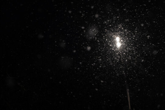 Falling snow particles illuminated by bright light source creating glowing blizzard effect against pitch black night background.