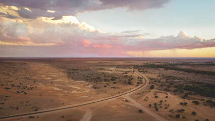 An aerial view of a winding road in a desert landscape at sunset, with a partly cloudy sky and rain in the distance.
