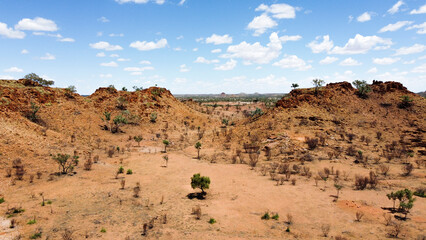 A low-angle aerial view of a dry, rocky desert landscape, featuring a valley between two reddish-brown hills with sparse trees and shrubs under a blue sky with scattered clouds. Near Mount Isa, QLD.