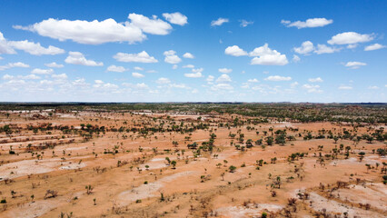 An aerial shot of a vast, flat, arid landscape dotted with scattered green trees and shrubs, extending to the horizon under a wide blue sky with fluffy white clouds. Near Mount Isa, QLD.