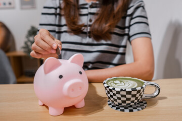 A woman places a coin into a pink piggy bank while enjoying a matcha latte, symbolizing savings and...
