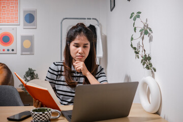 Young woman concentrating on work at home, using a laptop and notebook in a stylish, minimalist...