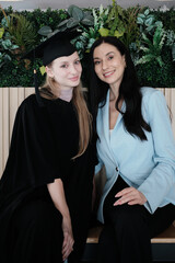 White mom and her graduate daughter sit and look at camera together with pride, love, in diploma...