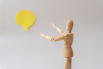 Small wooden mannequin pointing towards a yellow speech bubble in the background