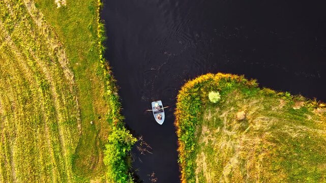 Top view of a rowing boat sailing on river surrounded by green grassy banks. Drone shot.
