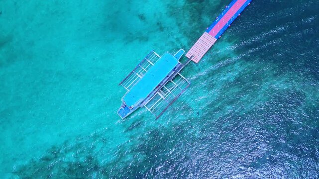 Spectacular 4K overhead drone shot of a traditional outrigger boat moored to a floating dock over crystal-clear turquoise waters in Boracay, Philippines.