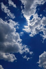 Obraz premium A vibrant, high-angle view of a partly cloudy sky. Bright sunlight peeks through fluffy white and grey clouds against a deep blue backdrop