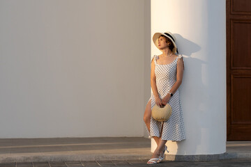 Woman wearing a white polka dot dress and black band straw hat gazes upward beside a white pavilion...