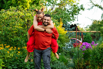 Fototapeta premium A cheerful man carries a woman on his back while standing in a lush garden surrounded by blooming flowers and greenery, capturing a moment of happiness and closeness