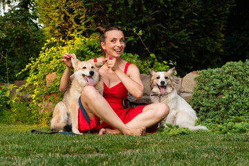 A cheerful woman in a red dress sits barefoot on green grass, playfully holding the ears of one...