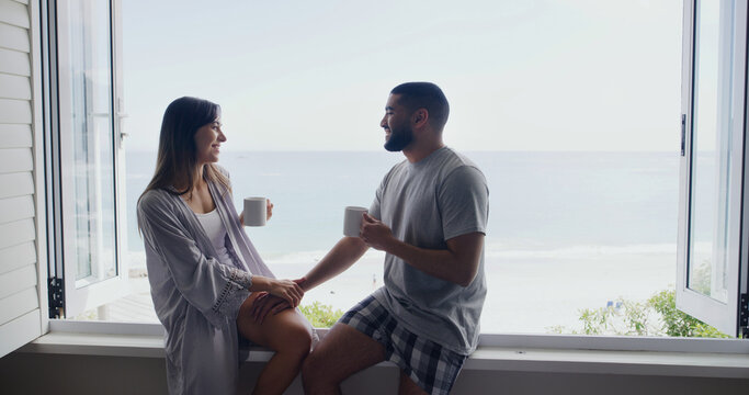Ocean, window and couple with coffee, home and holding hands with relationship, relax and discussion. Apartment, seaside and happy man with woman, talk or cappuccino with break, conversation and love - Powered by Adobe