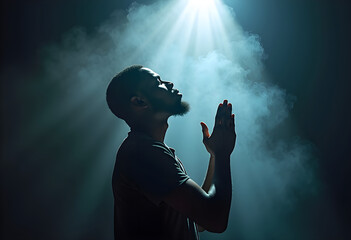 Young black man praying in the dark room with his hands folded in prayer, a beam of light shines on him from above