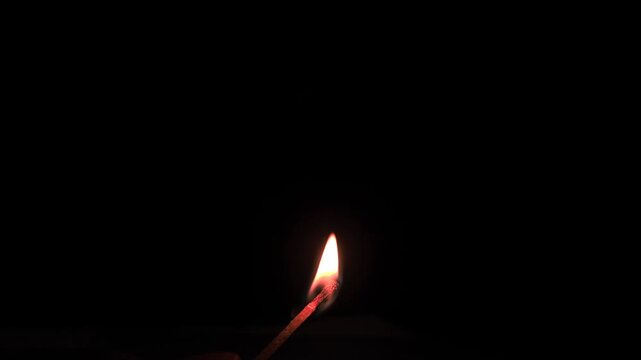 Extreme close-up captures fingers lighting a match, the sulfur tip igniting with fire and smoke, burning brightly in the darkness.