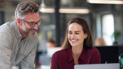 A middle-aged Caucasian man with glasses smiles at a young Hispanic woman in a modern office. They are engaged in a discussion over a laptop.