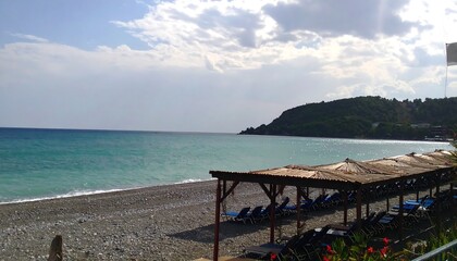 Serene coastal scene; pebble beach with sunshades and loungers, overlooking calm turquoise sea and verdant hills under a partly cloudy sky