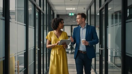 Two business people walking and discussing work in the office hallway, using a tablet and having a coffee, working together as a team - Powered by Adobe